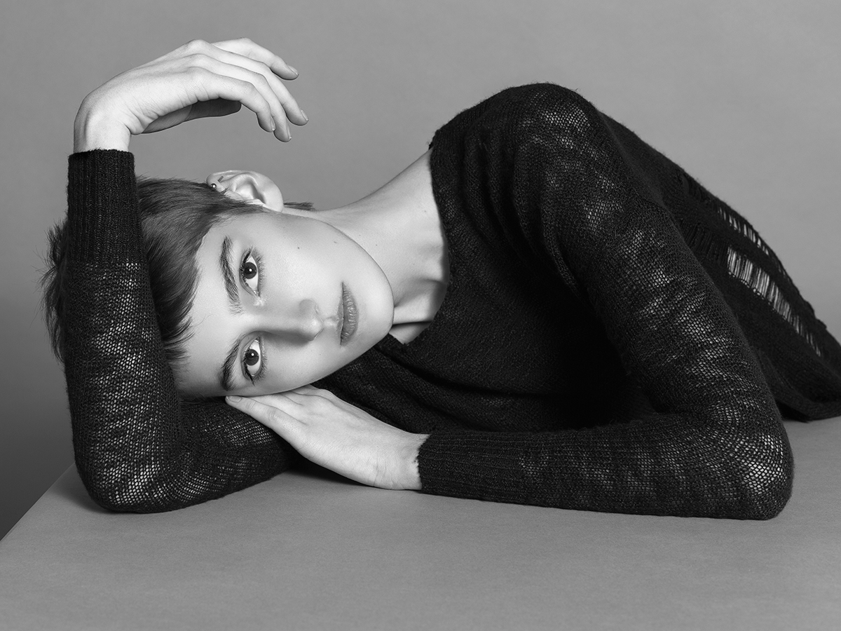 Model Kat McHenry poses on grey table surface in front of grey backdrop while wearing a sheer black top photographed by Joshua Pyles