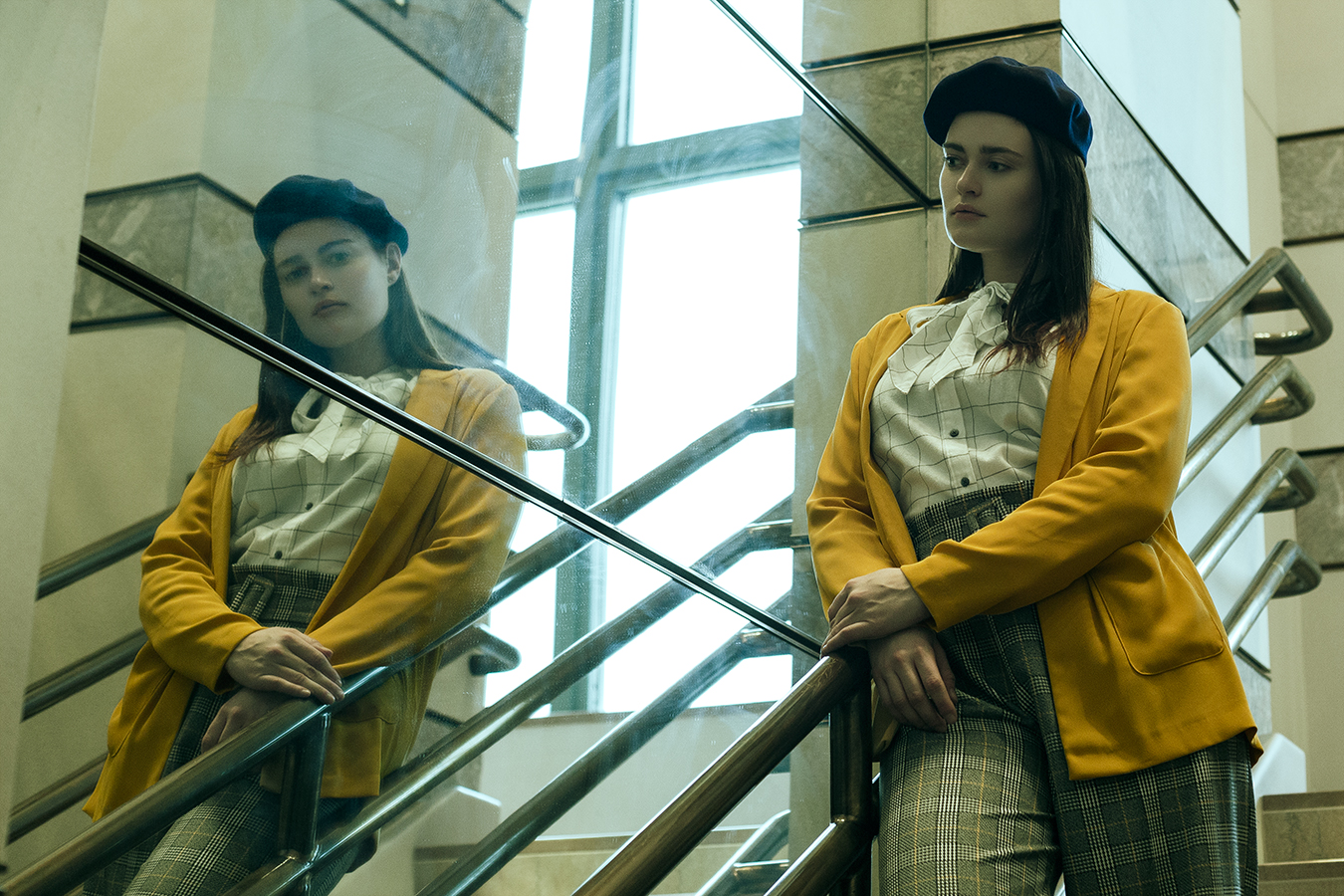 Model Malia Greiner looking at her reflection in a mirrored wall wearing plaid pants, checked blouse, yellow jacket, and beret photographed on location by Joshua Pyles